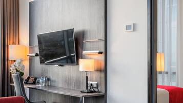 A modern hotel room with a flat-screen TV mounted on the wall, a desk with a lamp, a telephone, and a vase of flowers.