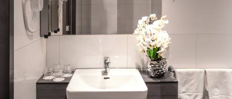Un baño con un lavamanos blanco, un espejo, un teléfono y un jarrón con flores blancas.