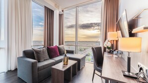 A modern hotel room with a large window, a sofa, a small table with a bottle and glass, a chair, and a desk with a lamp. - PLAZA Premium Almere (Almere)