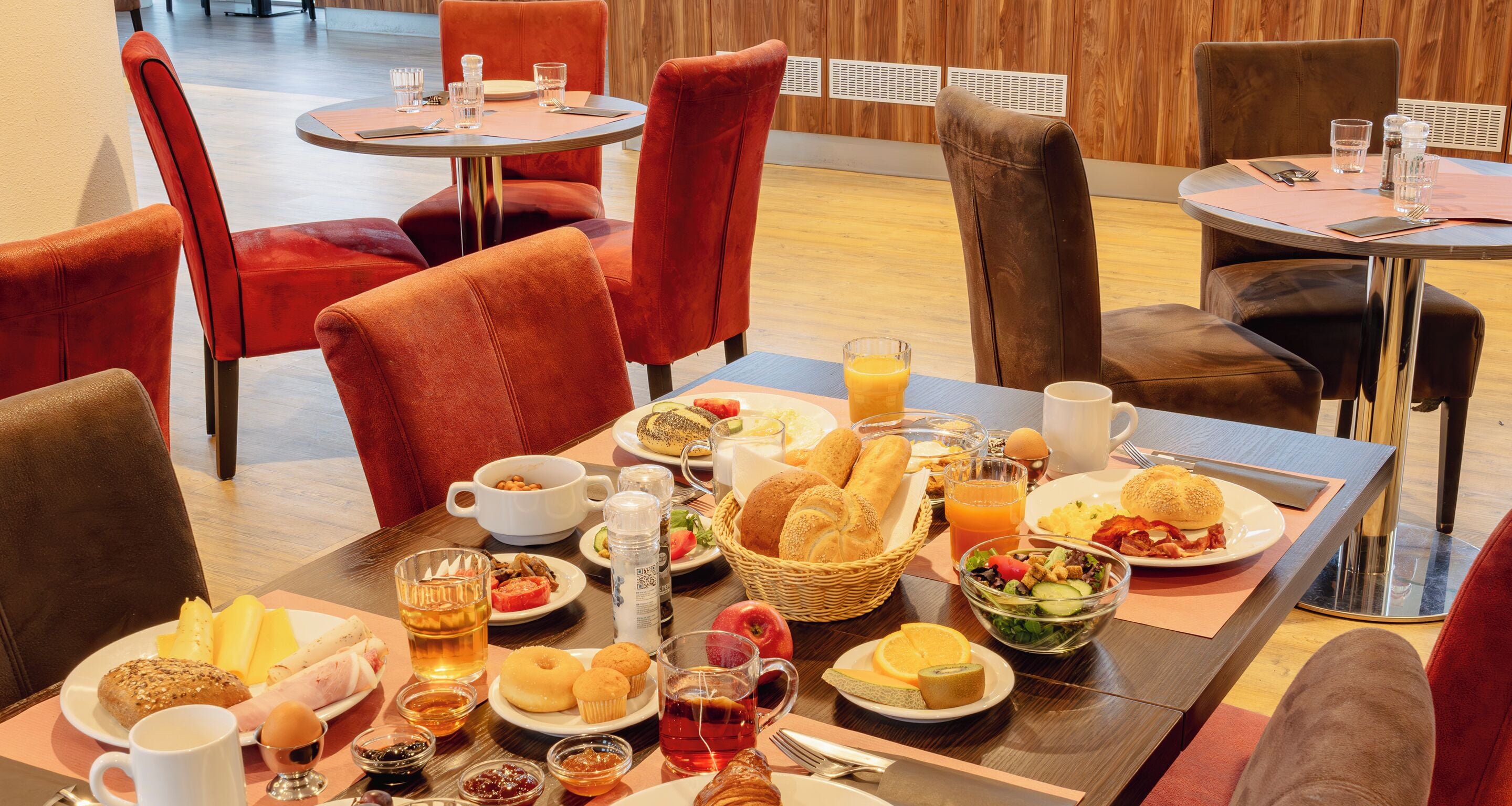 A breakfast buffet with various food items and beverages, arranged on a wooden table in a modern dining area with red upholstered chairs.