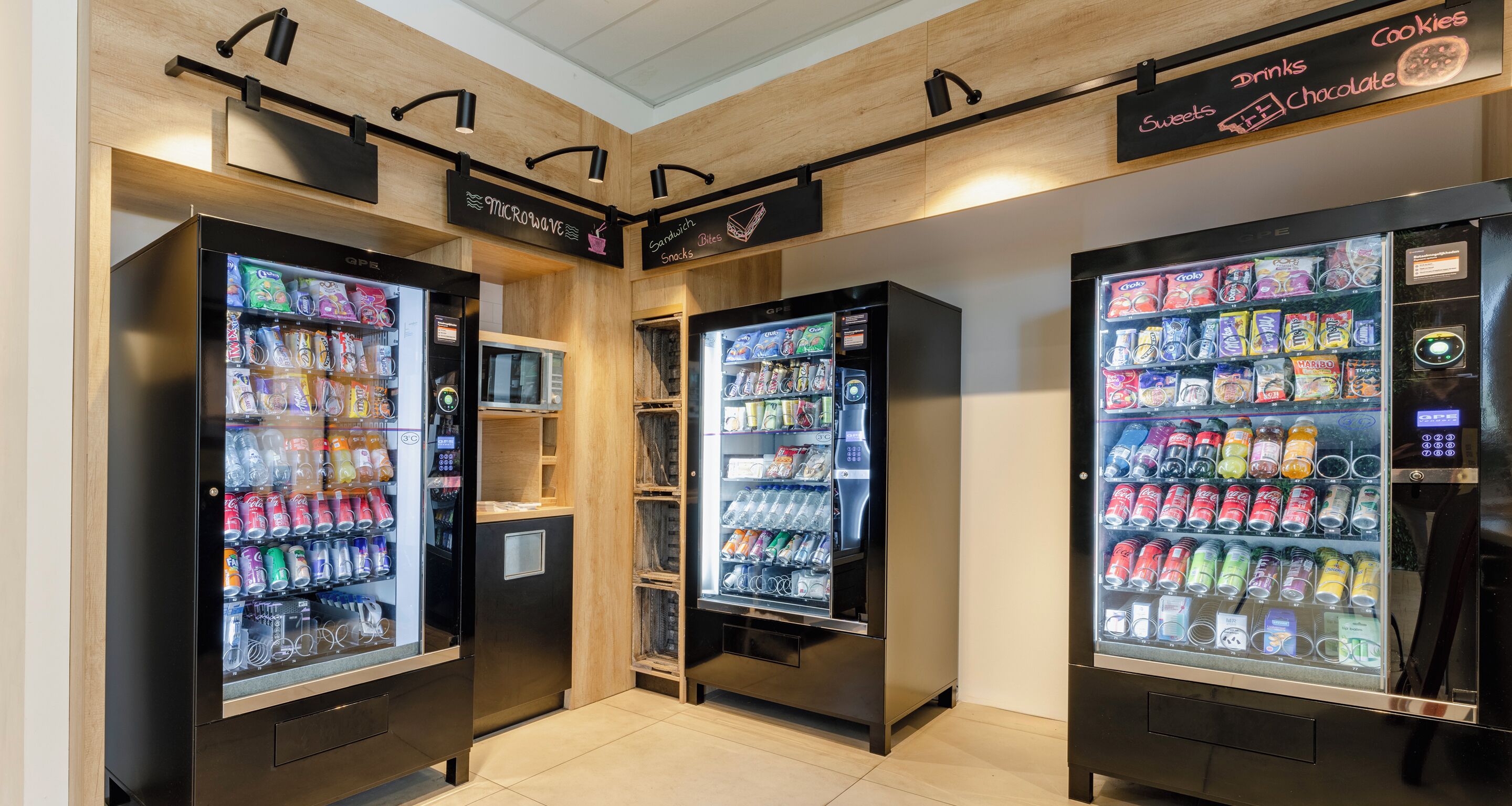 Three beverage vending machines with a variety of drinks and snacks, arranged in a row with wooden paneling and a ceiling light fixture.
