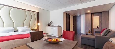 A hotel room with a bed, a dining table with fruit, a sofa, a kitchen area, and a bathroom.