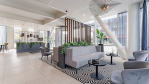 A modern lobby with a grey sofa, armchairs, a patterned rug, and a large window with a view of buildings. - PLAZA Premium Almere (Almere)