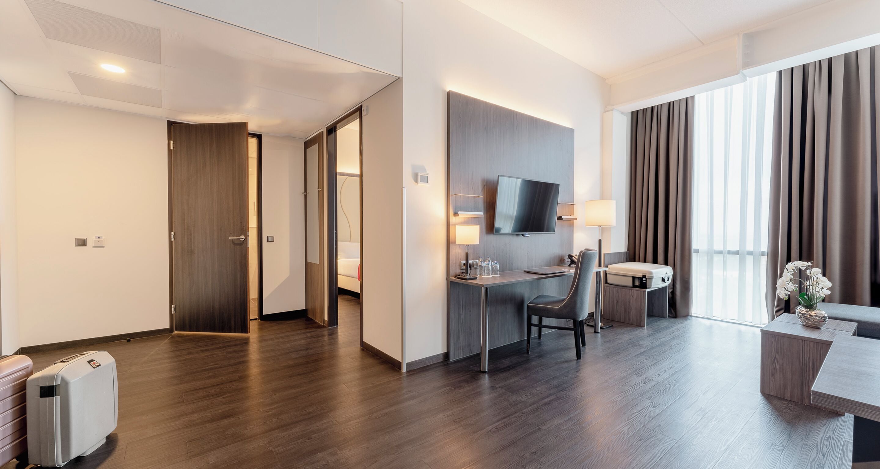 Een moderne hotelkamer met houten vloer, een bureau met stoel, een aan de muur bevestigde televisie, een nachtkastje met lamp en een groot raam met gordijnen.