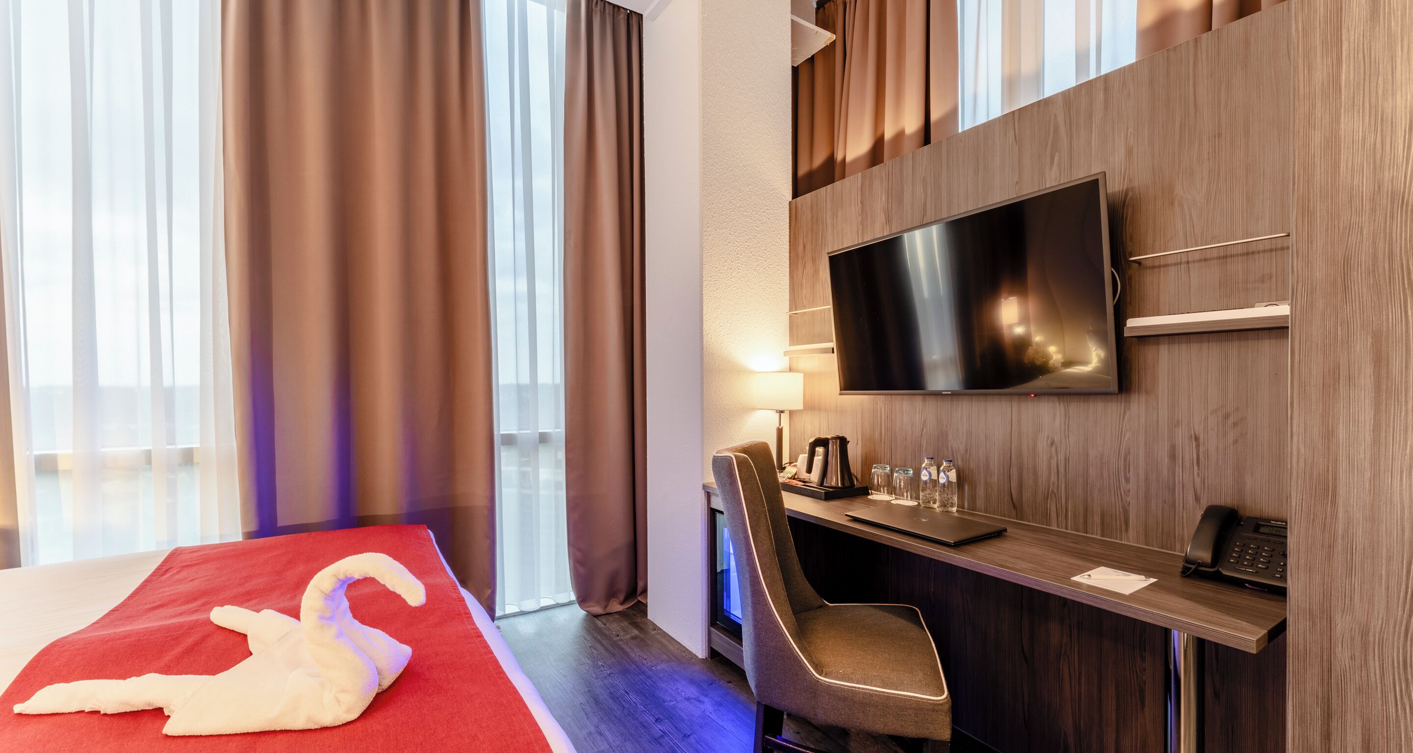 Een hotelkamer met een bed, een televisie, een bureau met stoel, en een raam met gordijnen.