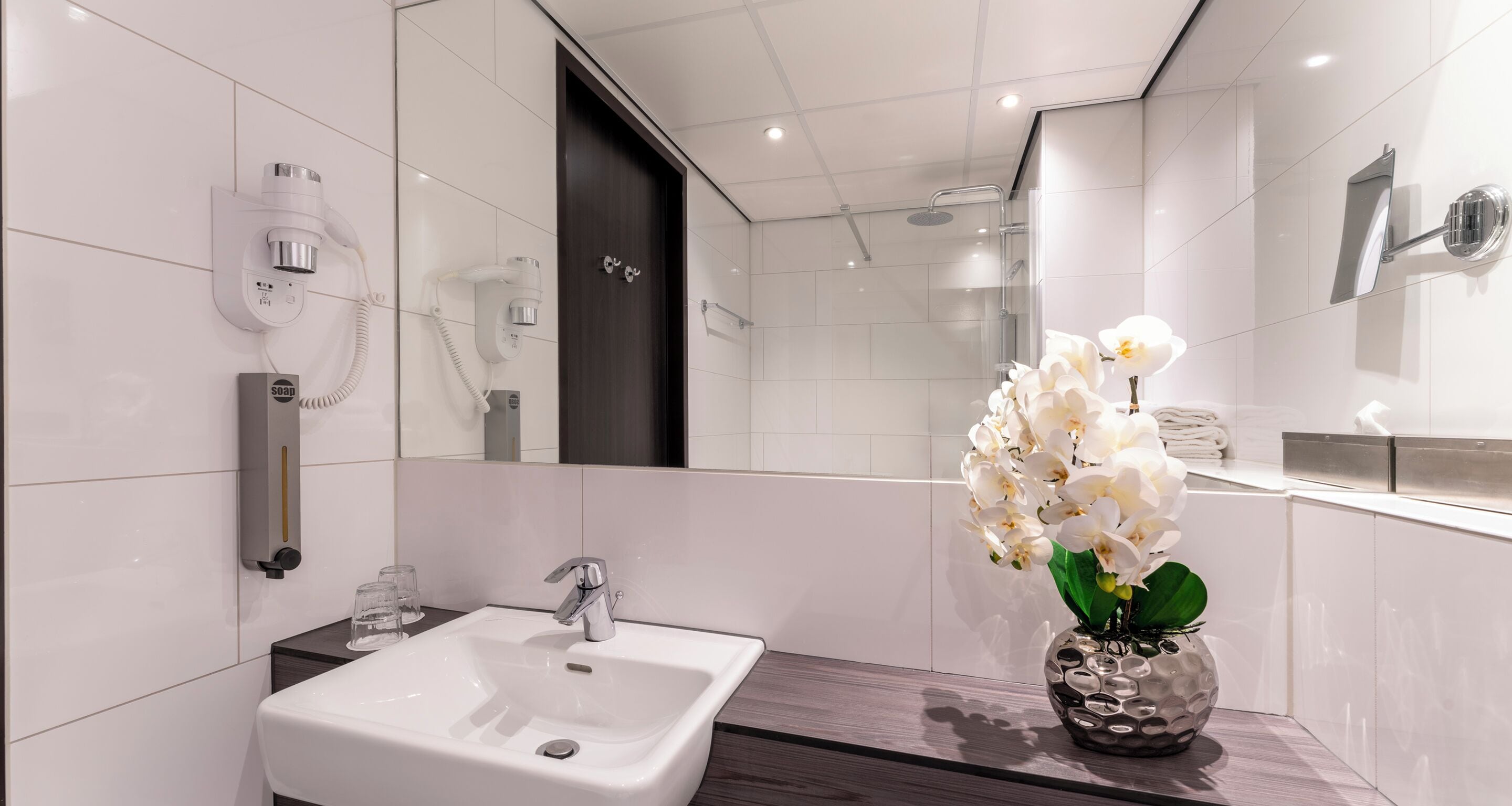 A modern bathroom with a white sink, a mirror, a shower, and a vase of white orchids.