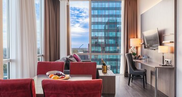Habitación de hotel con comedor, sofá, escritorio y un amplio ventanal con vista a la ciudad.