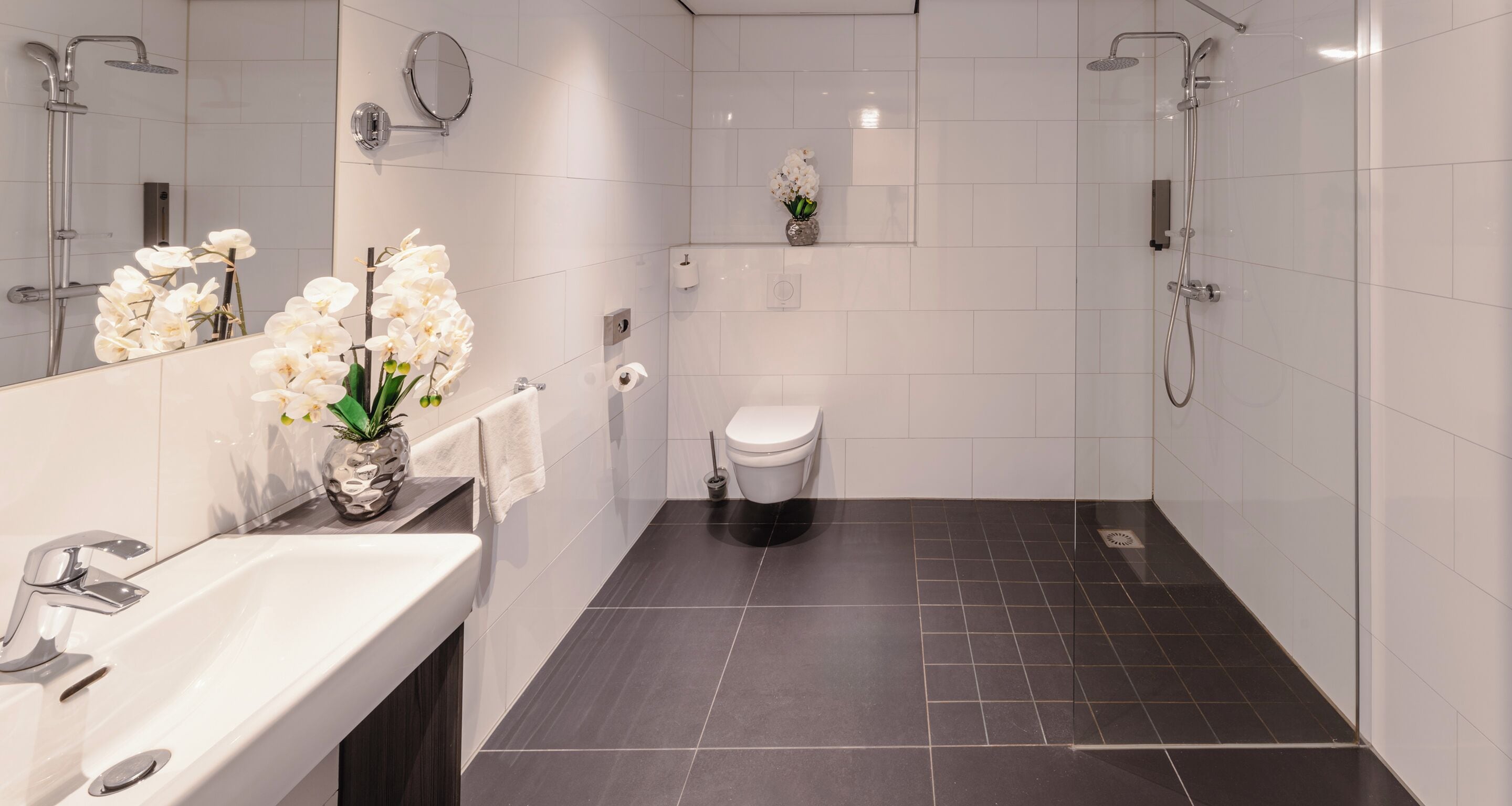 Une salle de bain moderne avec un lavabo, un miroir et des toilettes, le tout de couleur blanche. Un vase rempli de fleurs blanches est posé sur le comptoir du lavabo.