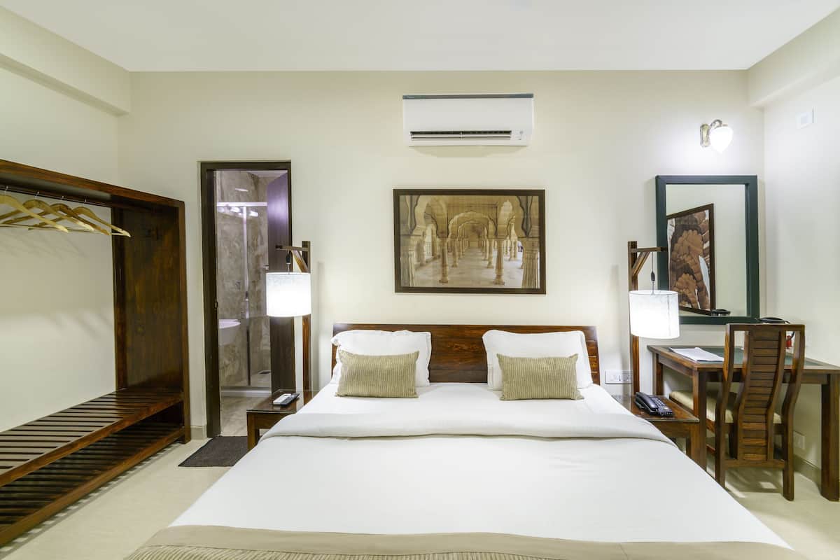 A hotel room with a large bed, a wooden wardrobe, a desk with a chair, a mirror, and a painting on the wall.