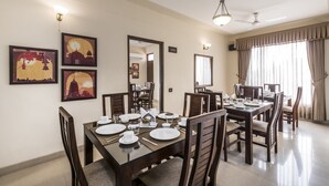 A dining area with a large table set for four, surrounded by wooden chairs. There are framed artworks on the wall, a ceiling fan, and a large window with curtains. - Zade House Jaipur (Jaipur)