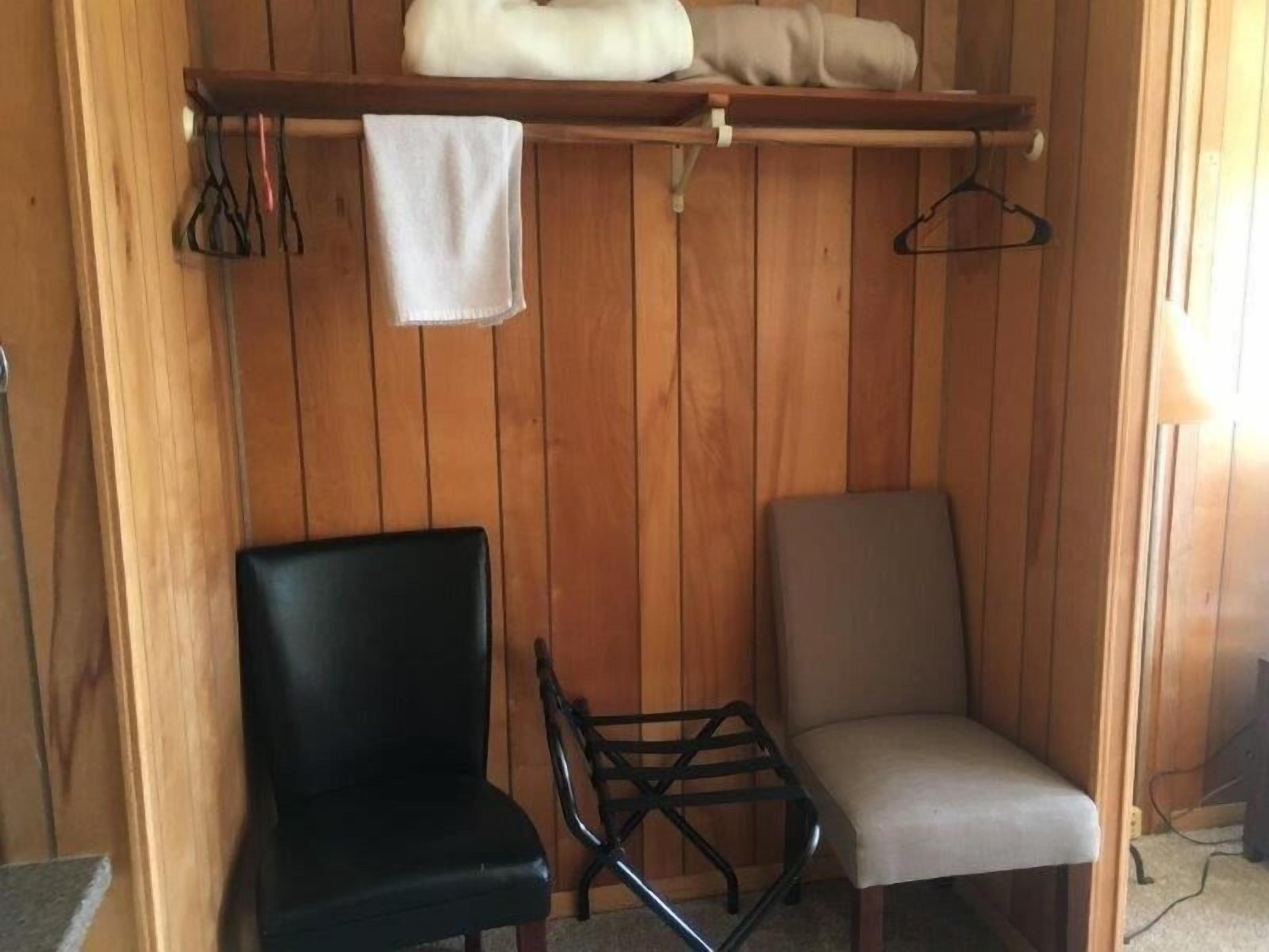 A room with wooden paneling, a black chair, a beige chair, and a shelf with a towel and clothes.