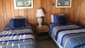 A room with two beds, each with blue bedding and decorative pillows. There are framed pictures on the wall, a lamp on a nightstand, and a small table between the beds. - Tyee Restaurant and Hotel (Coupeville)