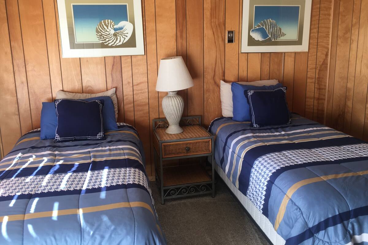 A room with two beds, each with blue bedding and decorative pillows. There are framed pictures on the wall, a lamp on a nightstand, and a small table between the beds.
