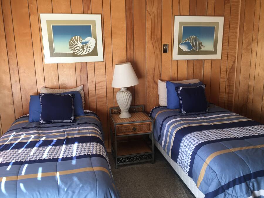 Une chambre avec deux lits, chacun agrémenté de draps bleus et de coussins décoratifs. Des tableaux encadrés ornent le mur, une lampe se trouve sur une table de chevet, et une petite table est placée entre les deux lits.