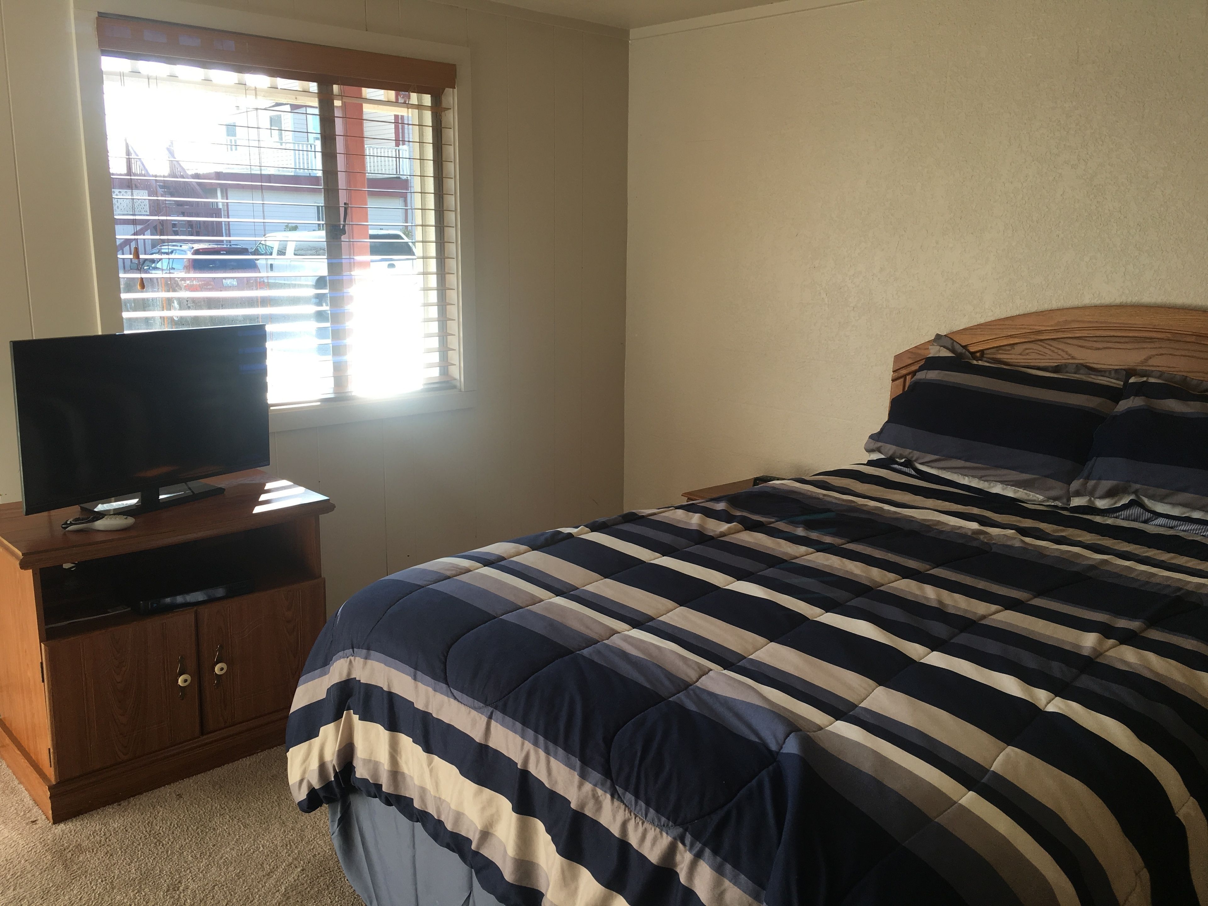A bedroom with a bed, a TV on a wooden stand, and a window with blinds.