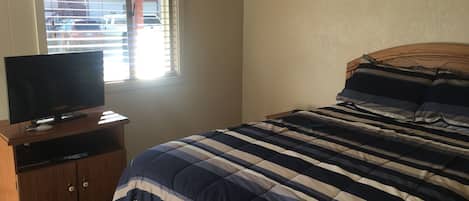 A bedroom with a bed, a TV on a wooden stand, and a window with blinds.