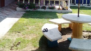 Un patio al aire libre con una mesa de piedra y bancos, rodeado de un jardín con flores y un toldo azul.