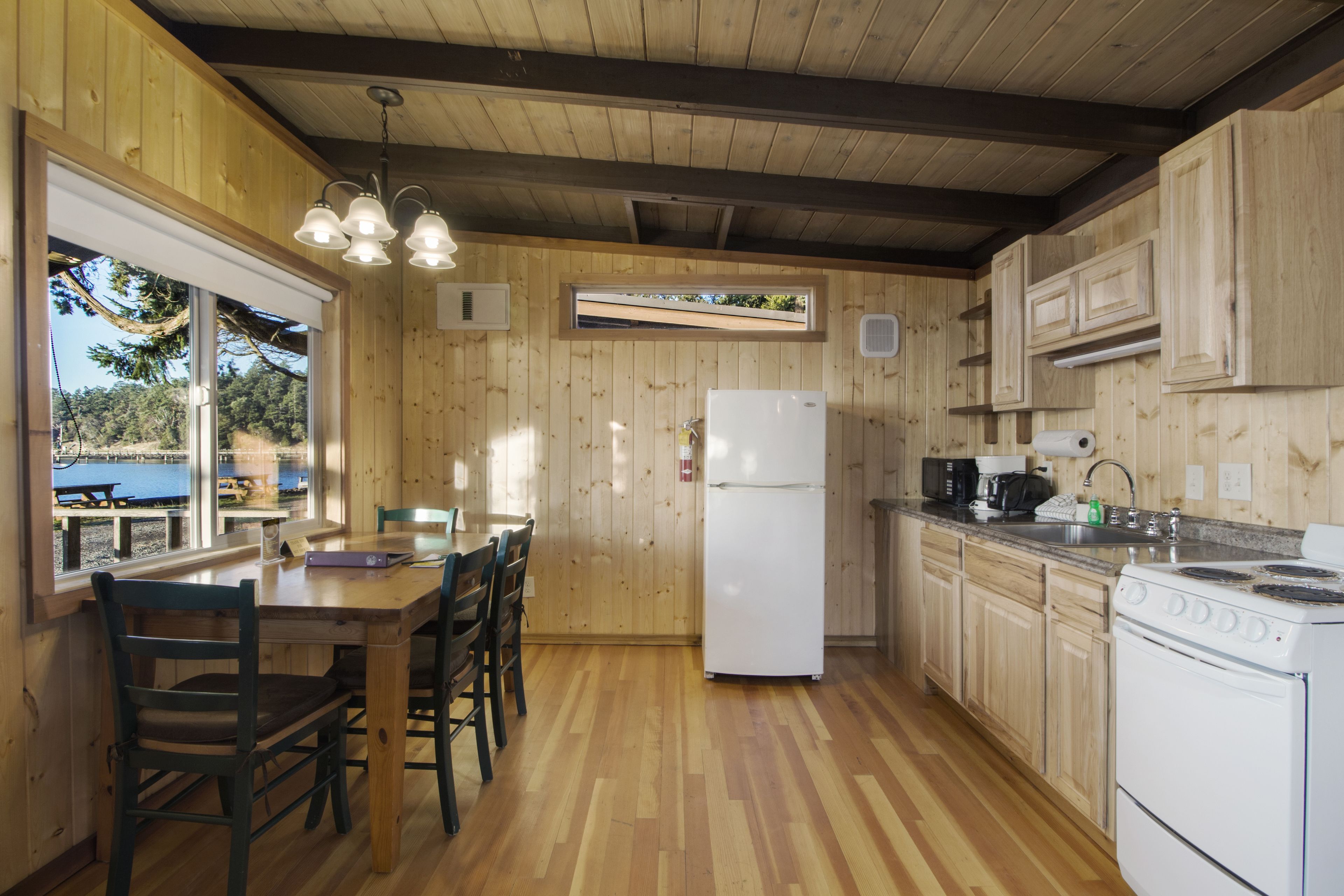 Une cuisine rustique aménagée dans une cabane en bois, avec une table à manger, des chaises, un réfrigérateur, une cuisinière et une fenêtre donnant sur un quai.