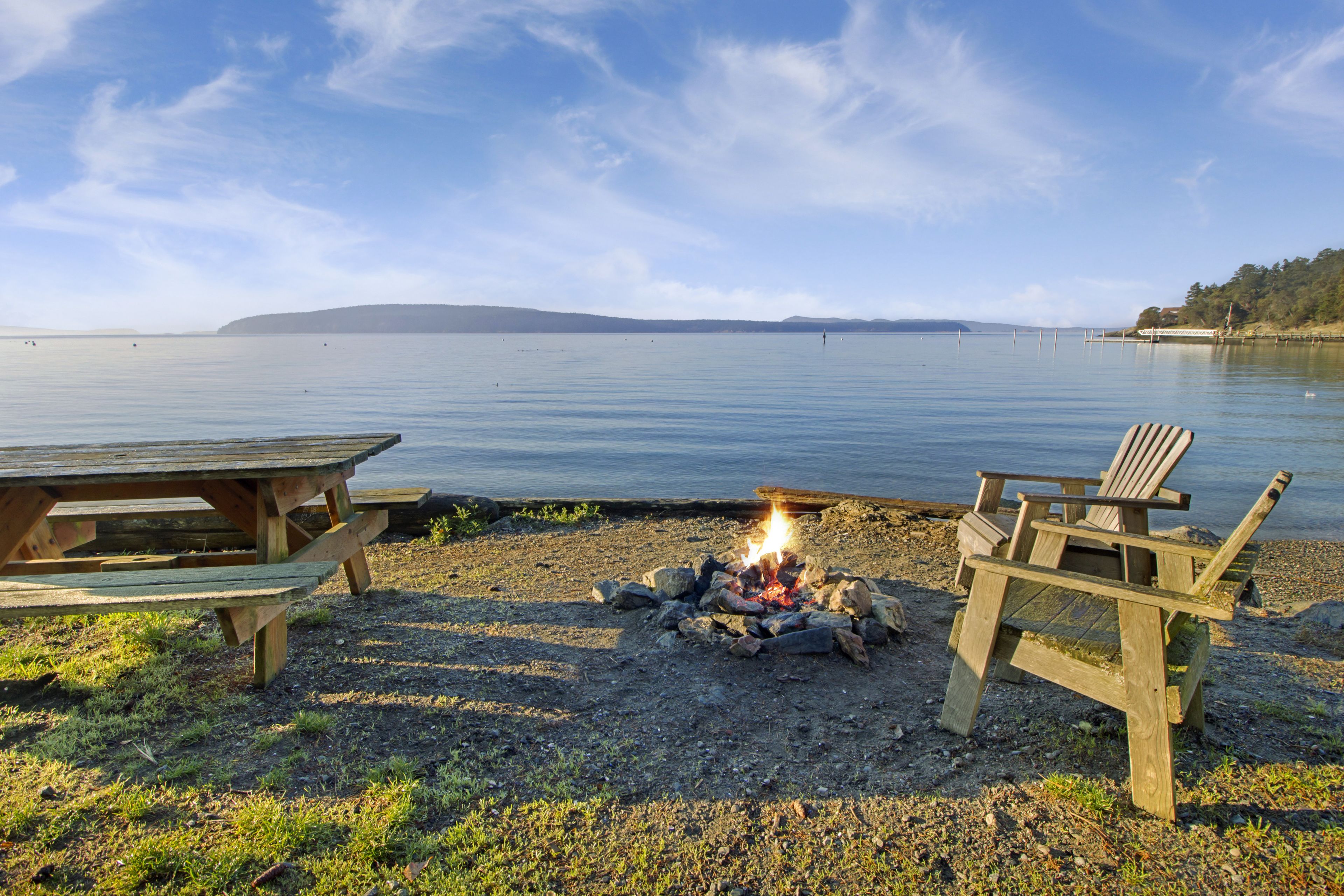 Un espace de pique-nique au bord du lac, équipé d’un foyer et de chaises en bois.