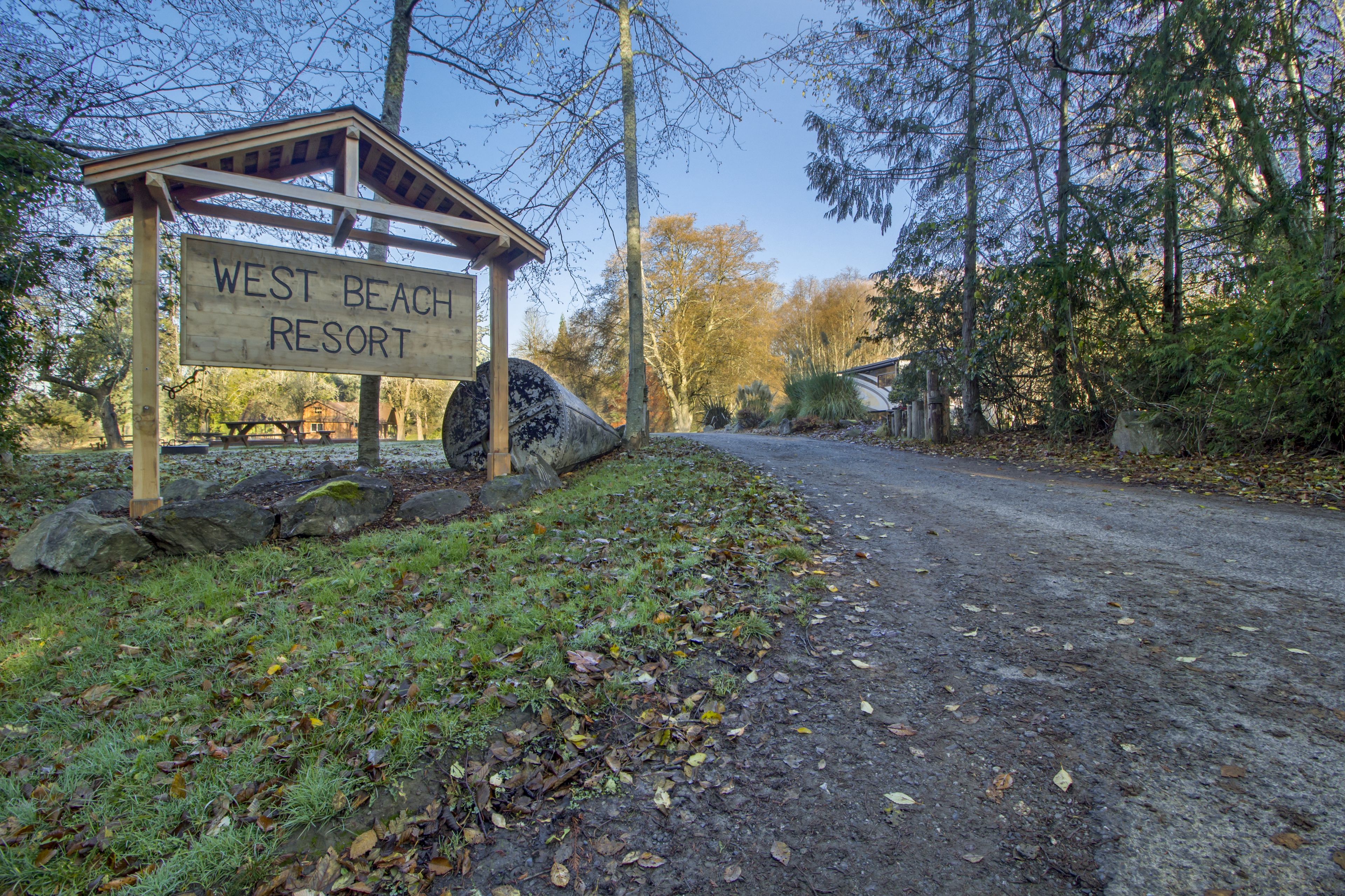 Ein Holzschild des West Beach Resort, mit einem Kiesweg, der darauf führt, umgeben von Bäumen und einem Picknicktisch.