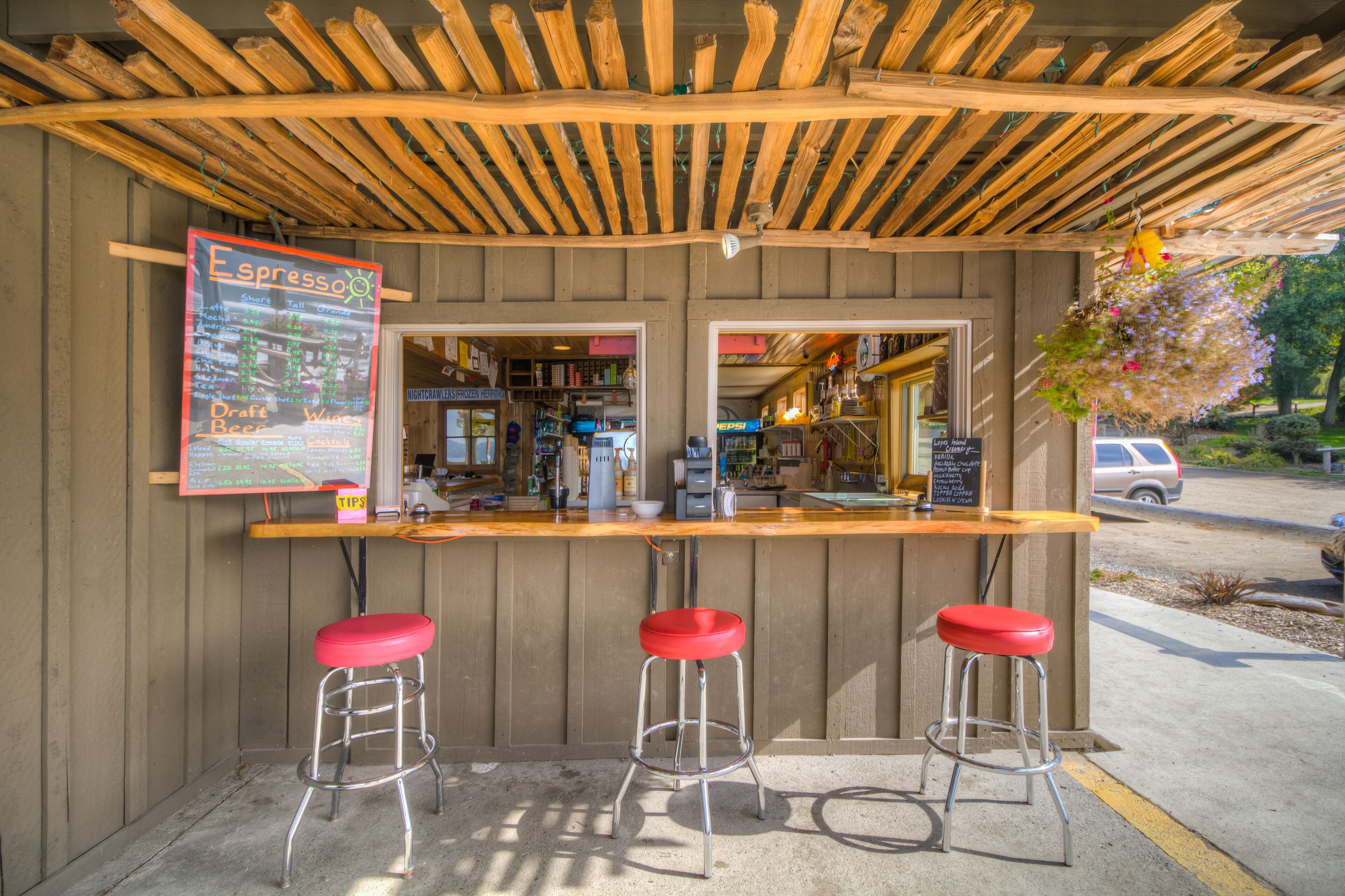 A small outdoor bar with a wooden pergola, a menu board, and two red stools.