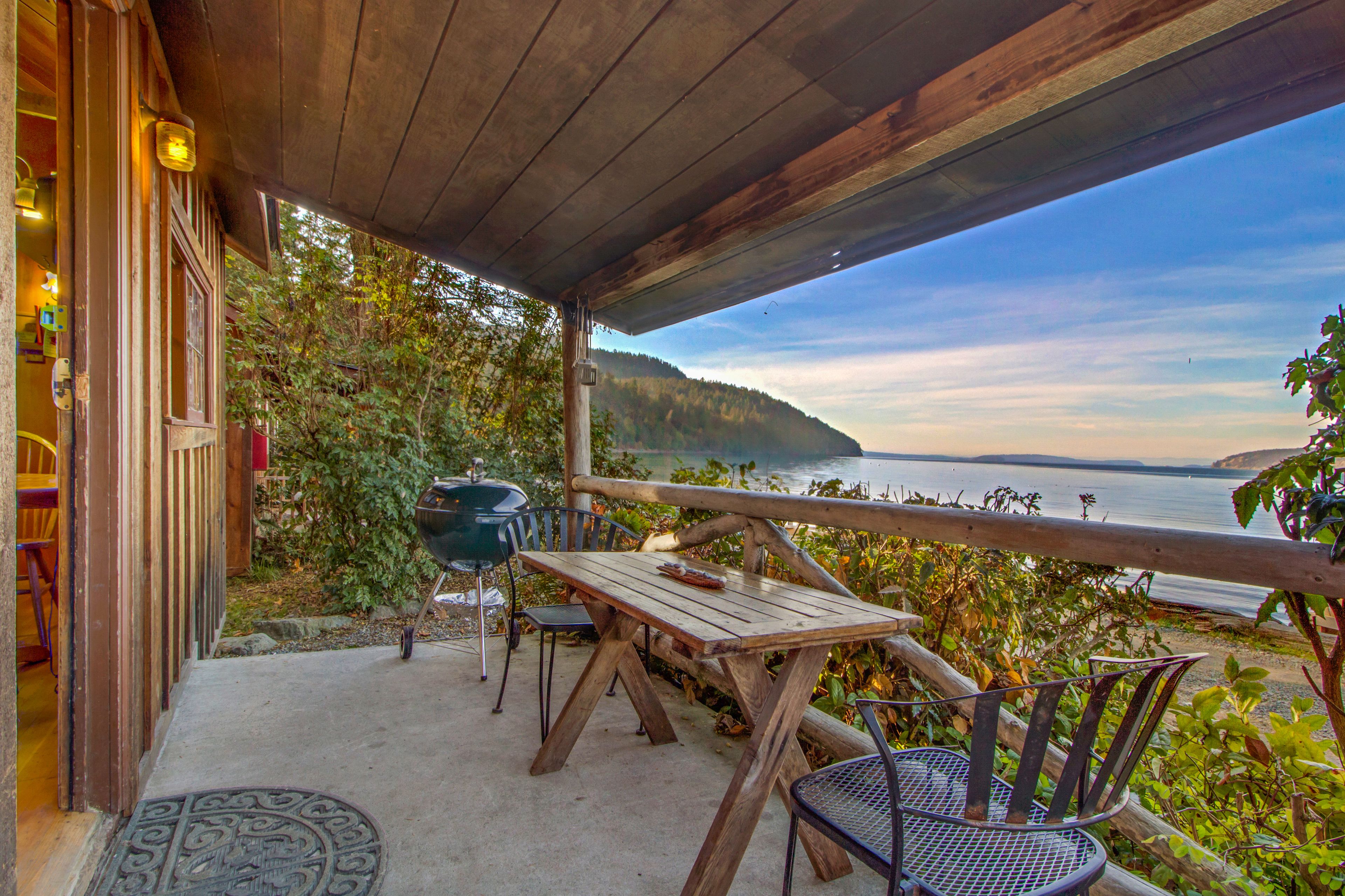 Une terrasse extérieure avec une table et des chaises en bois, offrant une vue sur une étendue d’eau et une colline boisée.