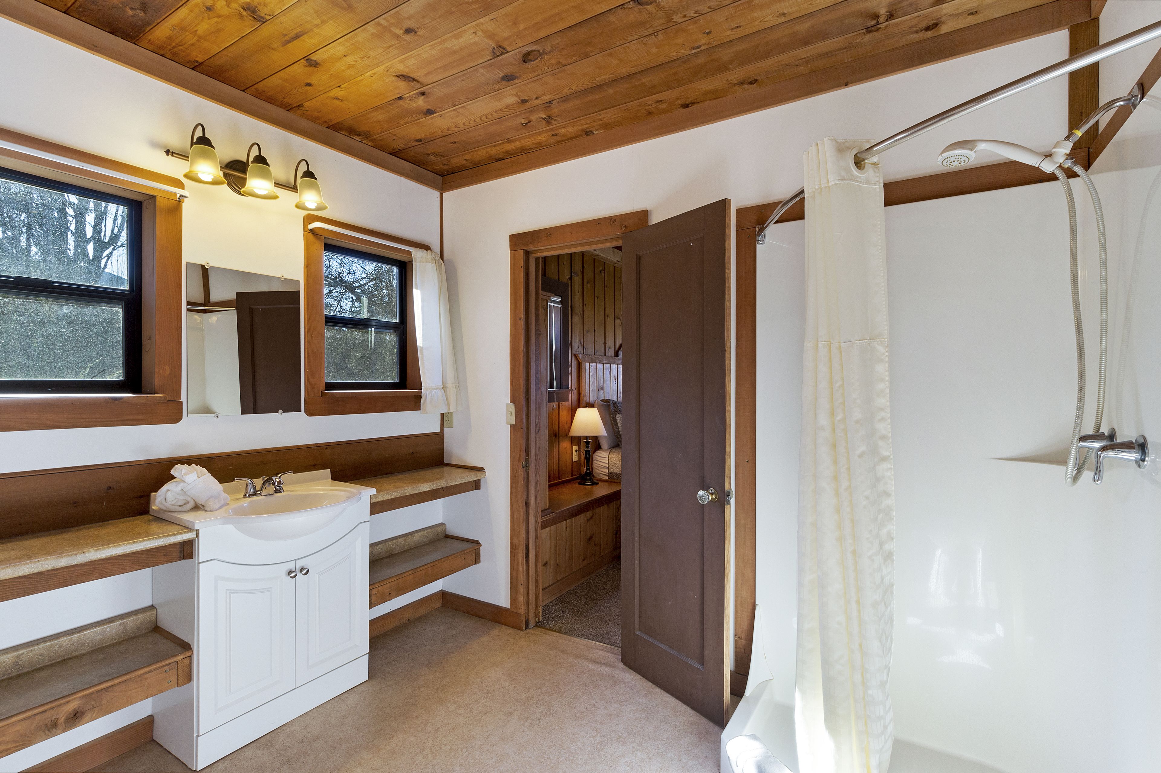 A bathroom with a wooden ceiling, a sink with a mirror, a shower with a curtain, and a wooden door leading to another room.