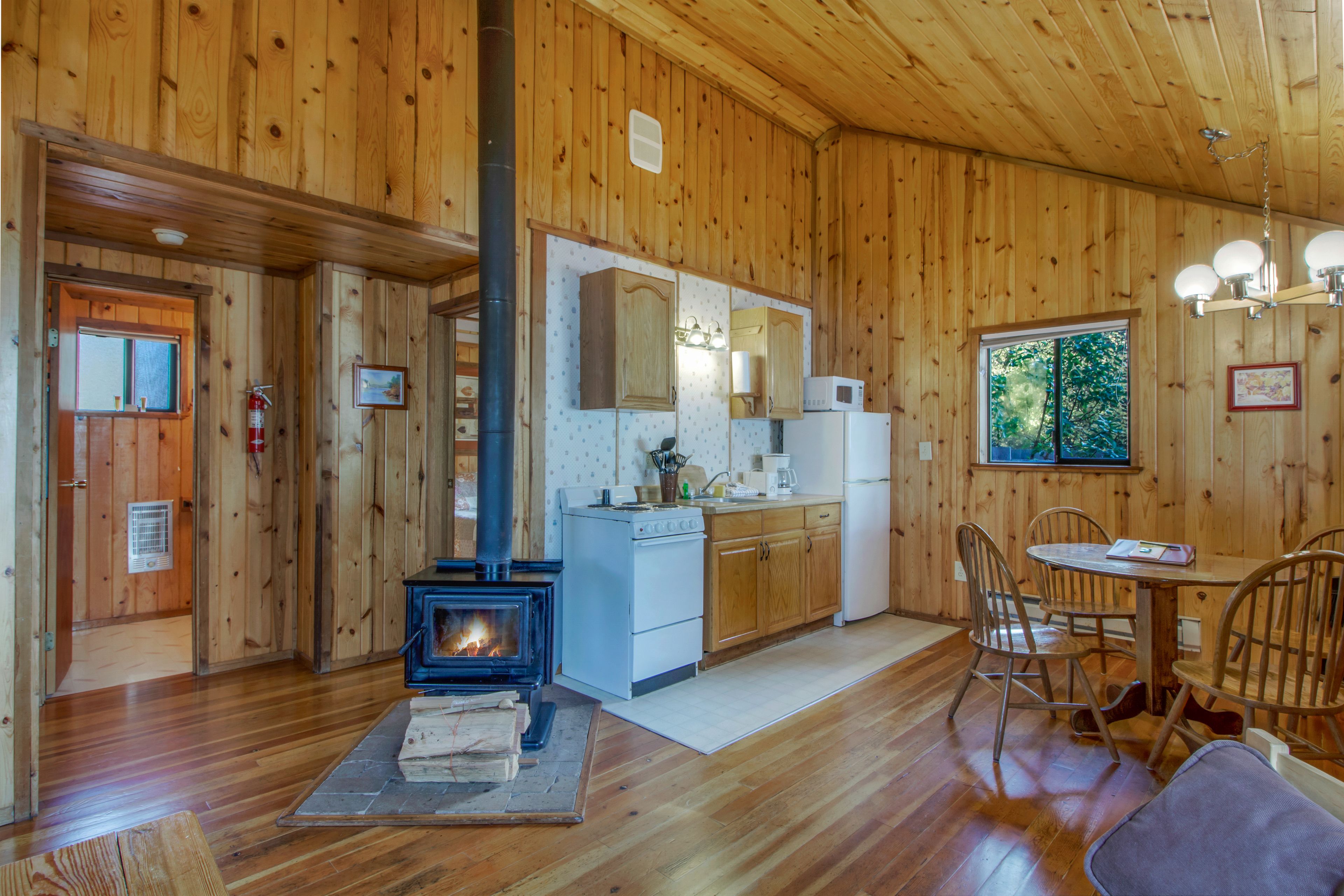 A wooden cabin interior with a wood-burning stove, kitchen area, dining table, and chairs.