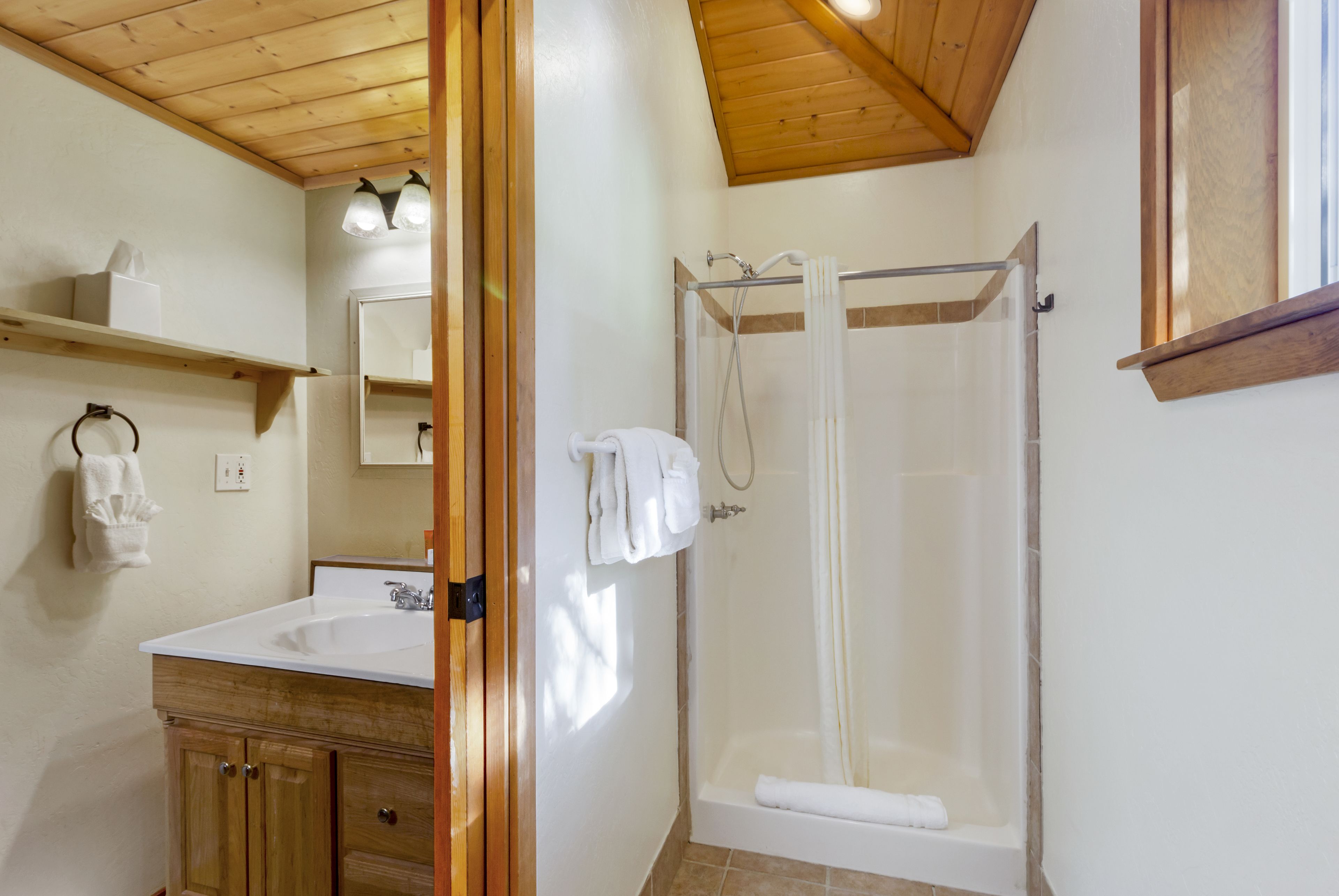 A bathroom with a wooden ceiling, a white sink, a glass shower enclosure, and a wooden cabinet.