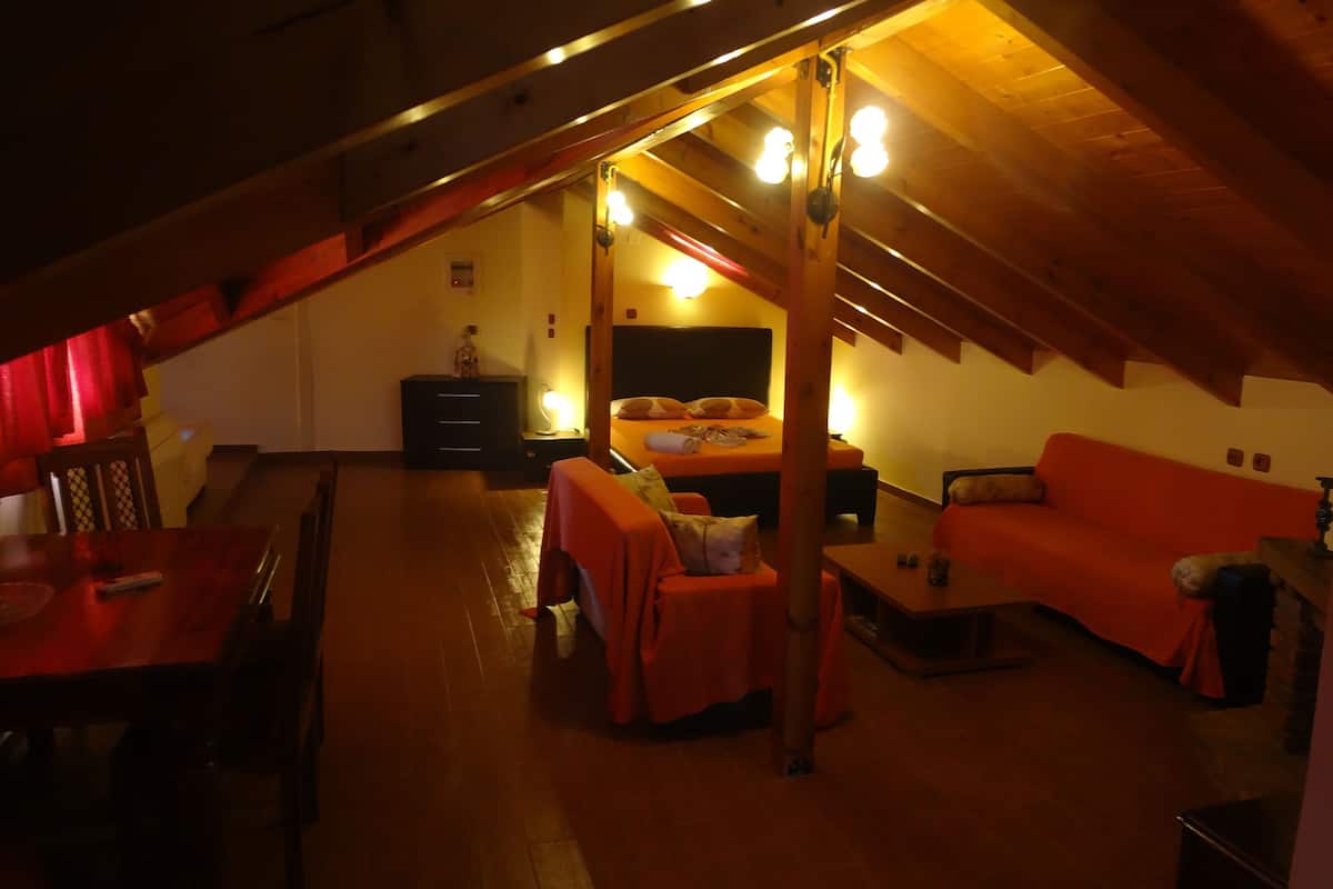 A cozy attic bedroom with a sloped ceiling, wooden beams, a bed, a sofa, and a dining table.