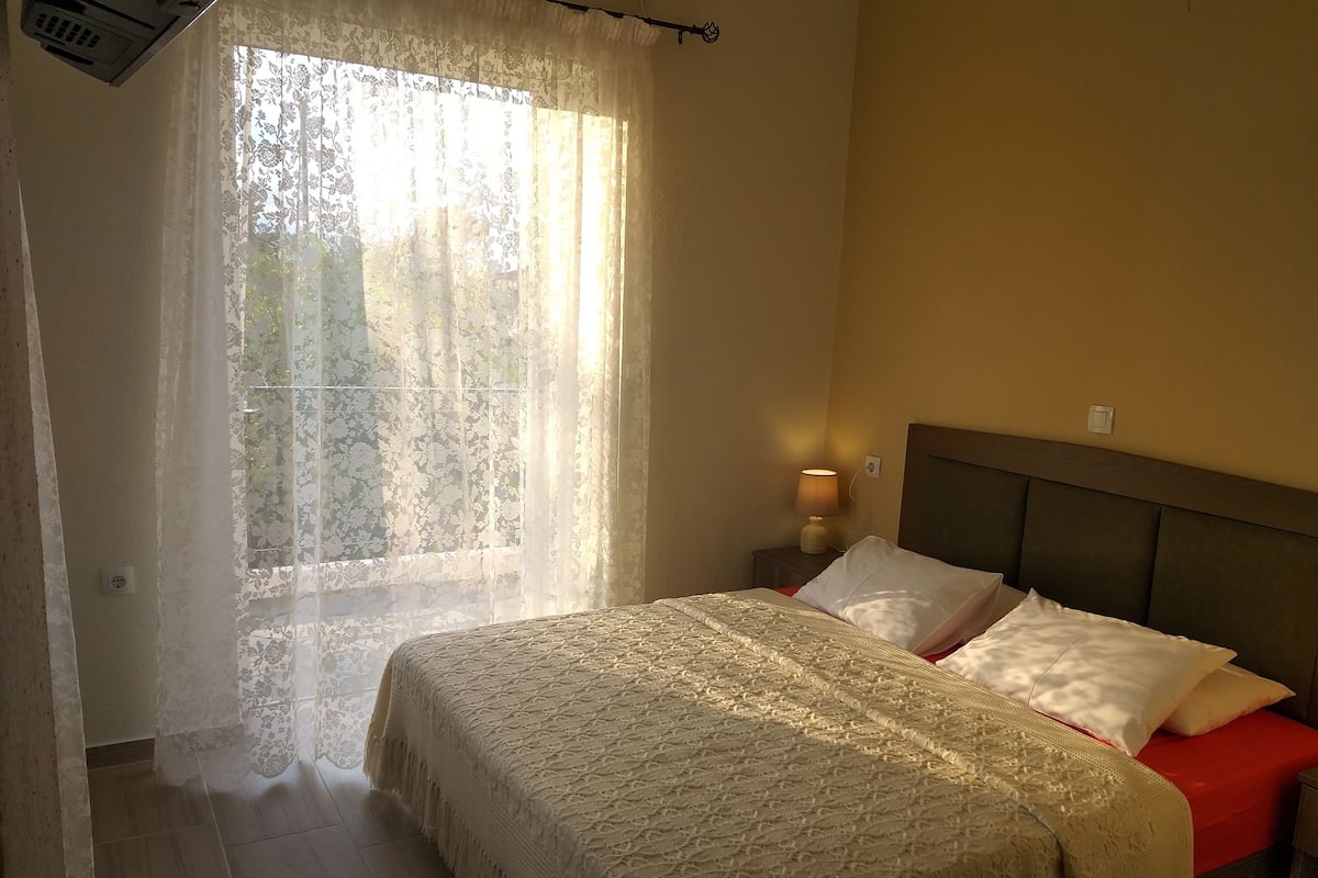 A bedroom with a bed, a nightstand, a lamp, and a window with sheer curtains.