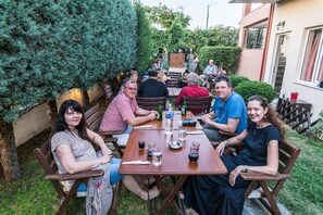 A group of people seated at an outdoor wooden table with drinks and a menu, surrounded by greenery and a building in the background. - Nostos Apartments (Tempi)