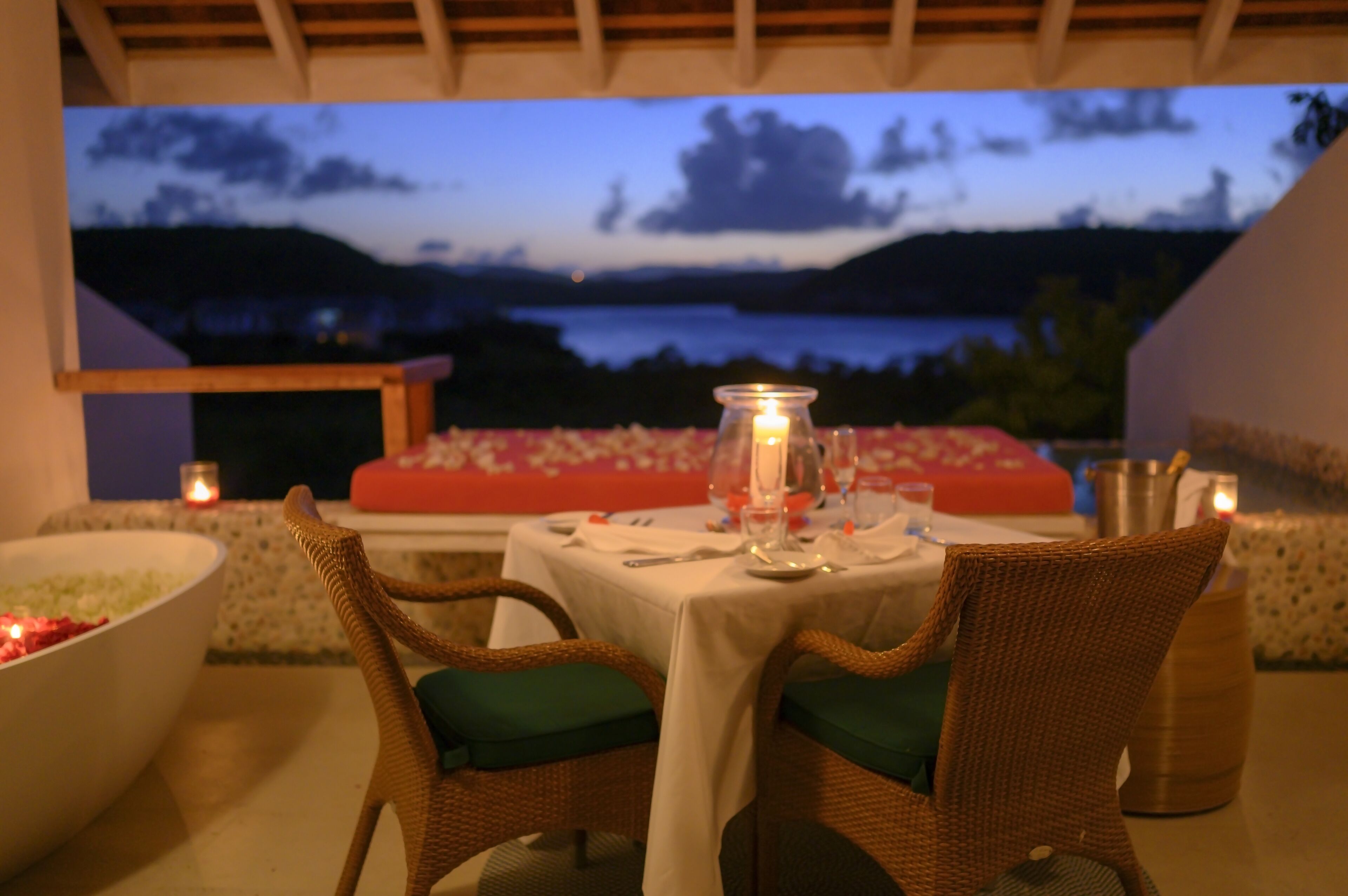 An outdoor dining area with wicker chairs and a table set for two, overlooking a scenic view of a lake and hills at dusk.