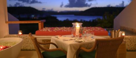An outdoor dining area with wicker chairs and a table set for two, overlooking a scenic view of a lake and hills at dusk.