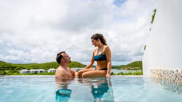 Un couple se trouve dans une piscine à eau claire, d’où l’on peut admirer un paysage verdoyant et luxuriant ainsi que des bâtiments situés au loin.