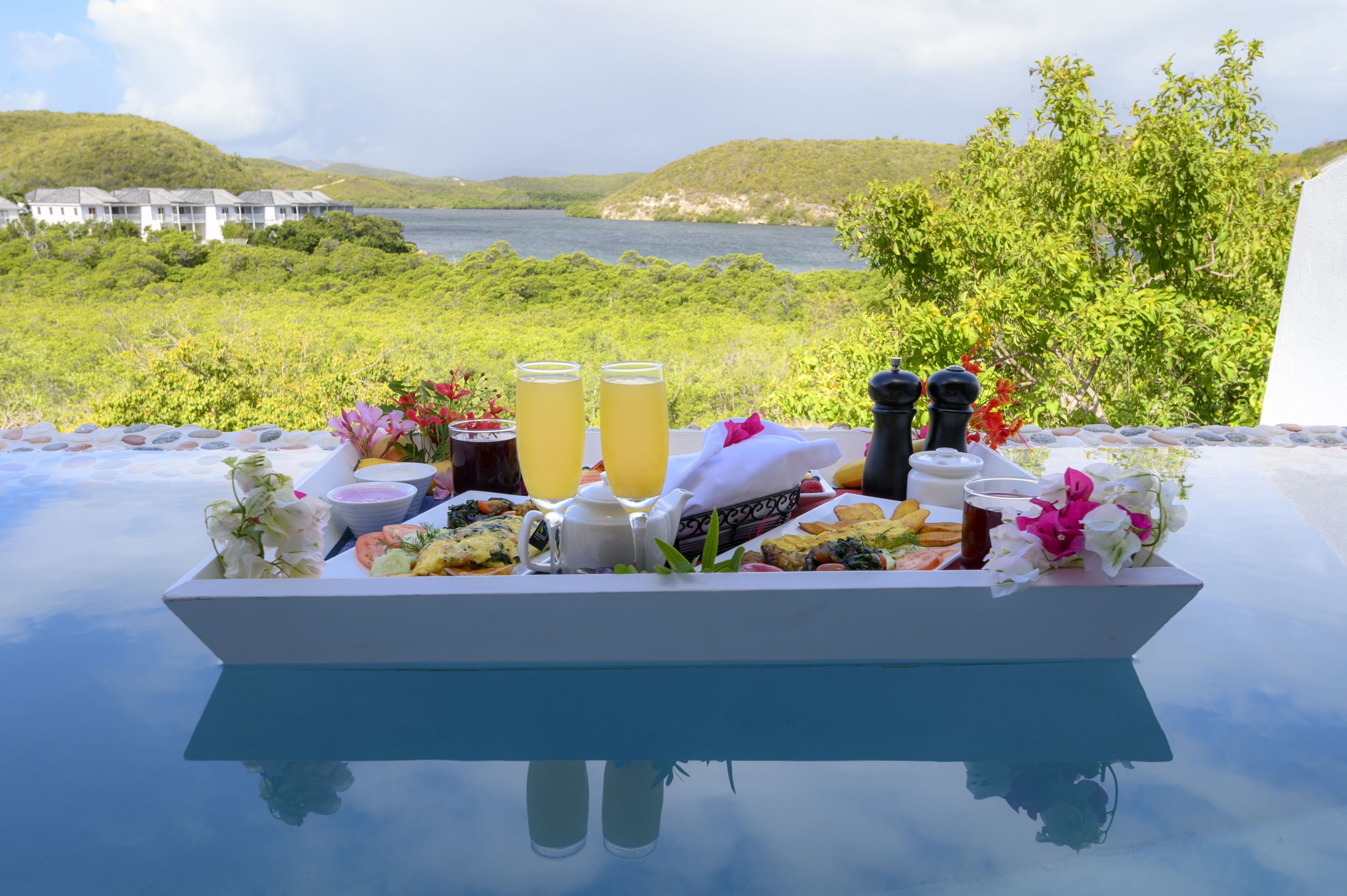 Bandeja de desayuno con bebidas y comida sobre una mesa de vidrio, con vista a un lago y colinas.