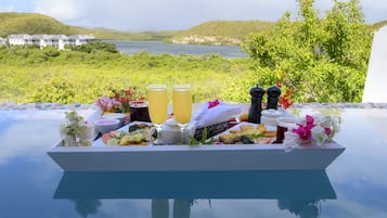 Bandeja de desayuno con bebidas y comida sobre una mesa de vidrio, con vista a un lago y colinas.