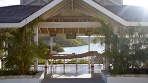 A covered outdoor area with a view of a lake, featuring a wooden structure with a white roof and columns, surrounded by plants and a rope barrier.