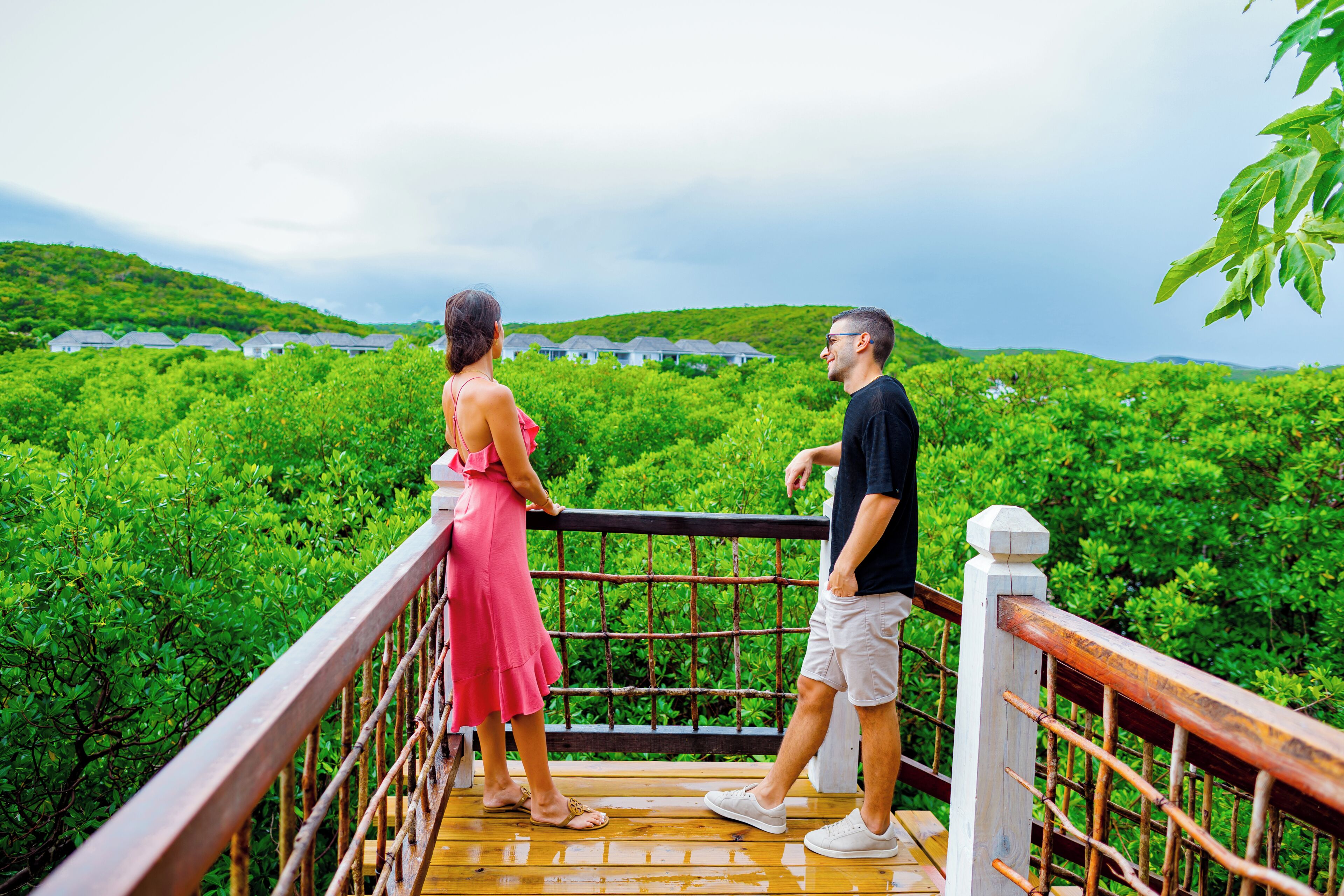 Una pareja parada sobre una terraza de madera con vistas a exuberante vegetación y casas al fondo.