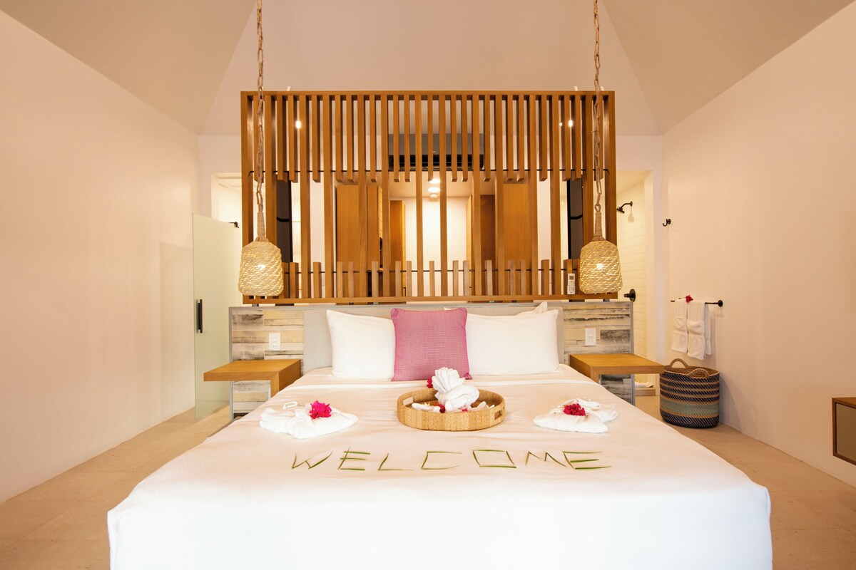 A hotel room with a large bed, a wooden headboard, a "WELCOME" sign on the bed, and a basket with towels.