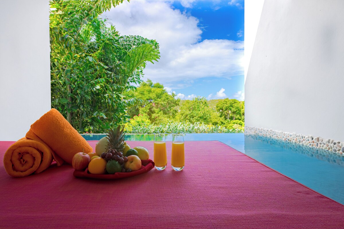 A red mat with a fruit bowl, two glasses of orange juice, and a rolled orange towel on a balcony overlooking a pool.