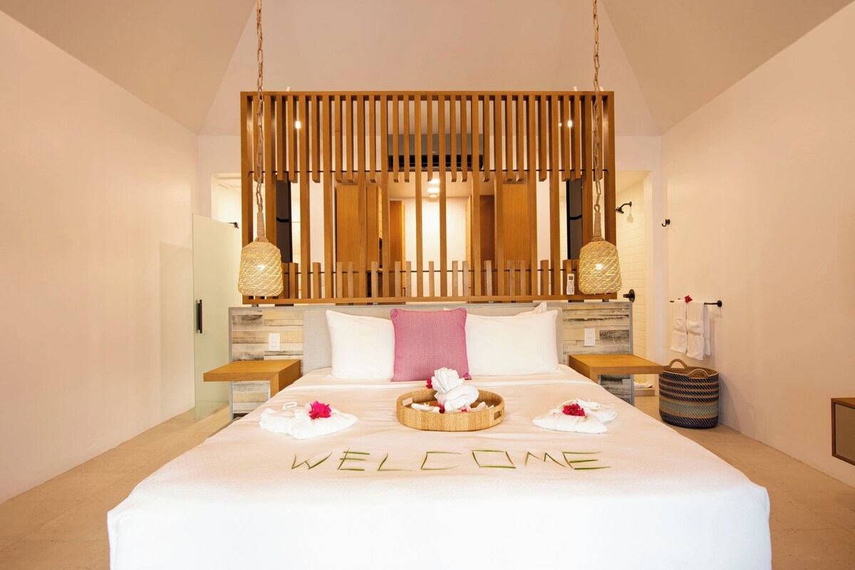 Habitación de hotel con una cama grande, un cartel que dice 'WELCOME', cabecera de madera con listones, dos lámparas de noche y una mesita de noche pequeña.
