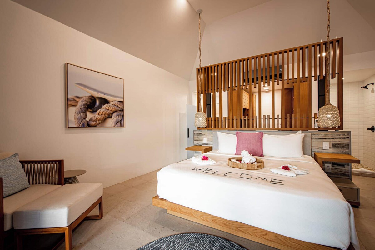 Een moderne hotelkamer met een bed, een stoel, een houten scheidingswand, een hanglamp en een lijst met een foto aan de muur.