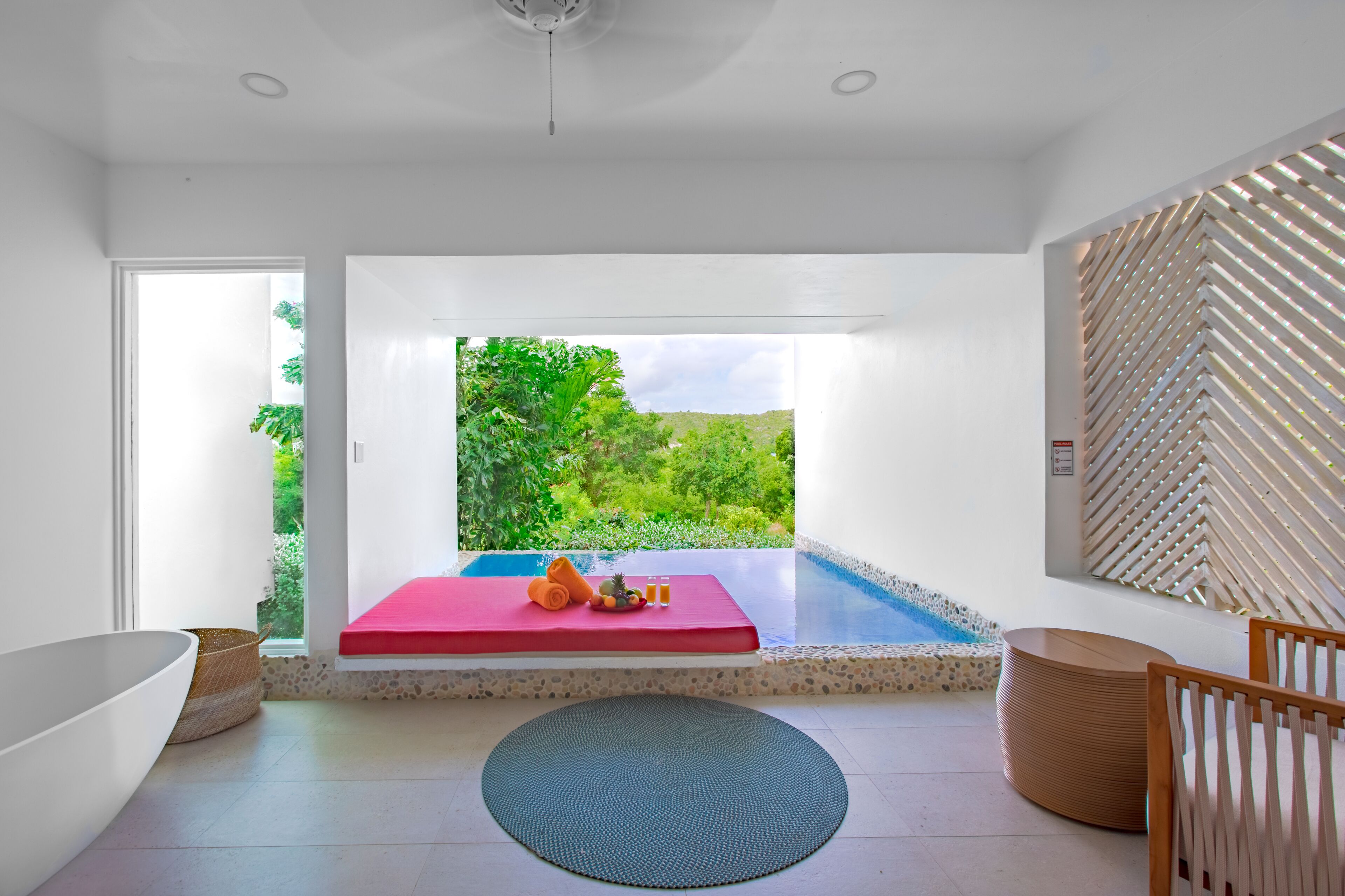 Une salle de bain moderne avec une grande baignoire, un banc rouge et une vue sur un jardin luxuriant, visible à travers une porte vitrée.