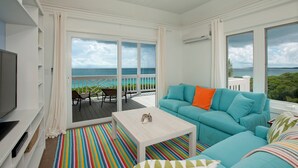 A bright room with a large window overlooking the sea, a teal sofa, and a colorful striped rug.
