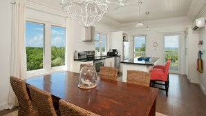 A spacious kitchen with large windows, a dining area with a wooden table and wicker chairs, and a view of the outdoors.