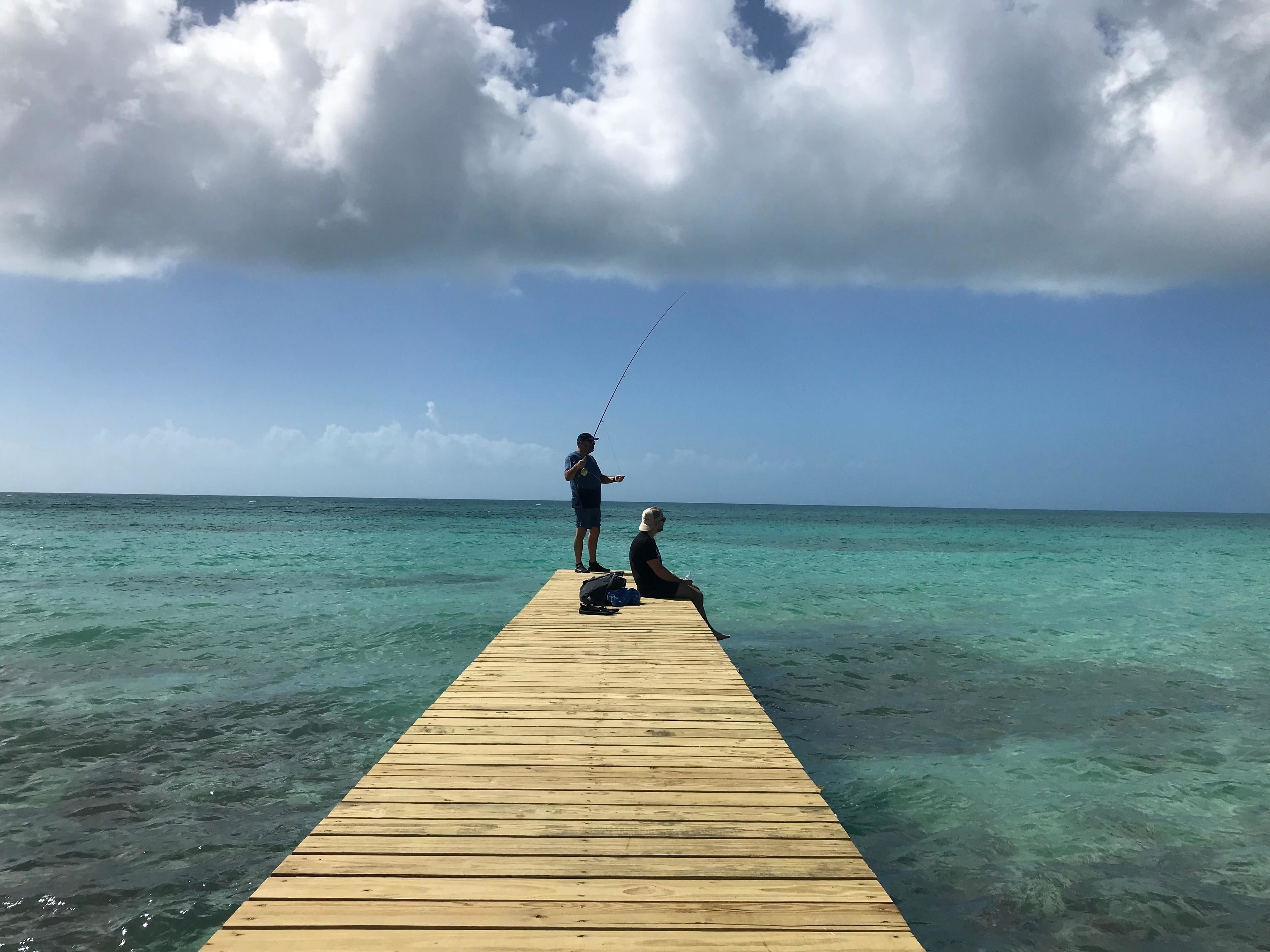 A wooden pier extending into clear turquoise waters with two people fishing.