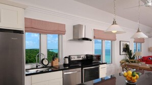 Une cuisine moderne équipée d’un réfrigérateur en acier inoxydable, d’une cuisinière et d’un four à micro-ondes. Une grande fenêtre offre une vue sur l’océan.