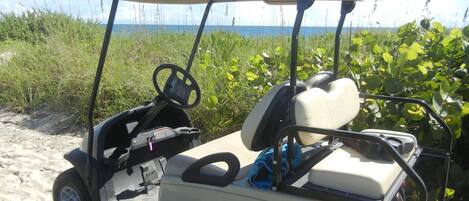 A golf cart parked on a sandy area with a view of the ocean and greenery in the background.
