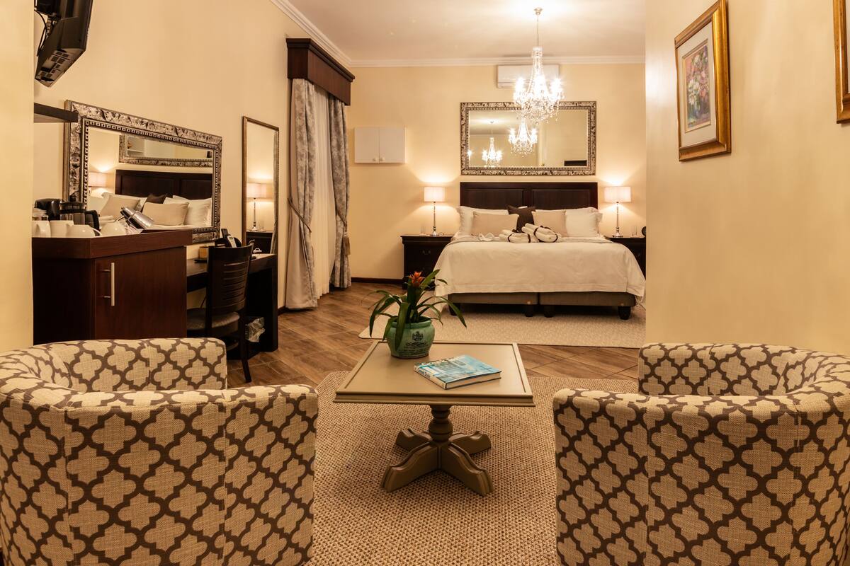 A hotel room with a bed, two armchairs, a small table, a desk with a chair, a mirror, a chandelier, and a potted plant.
